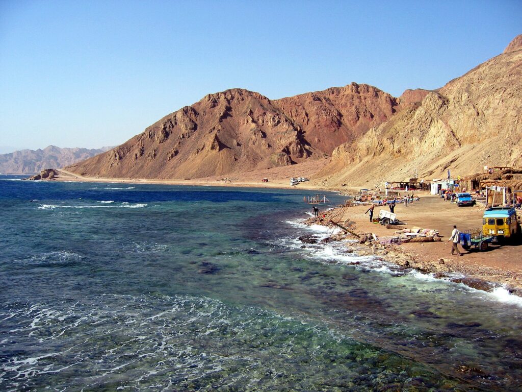 The Blue Hole beach: Bedouin cafés at the foot of ocher mountains, a few cars, rough sea in the foreground