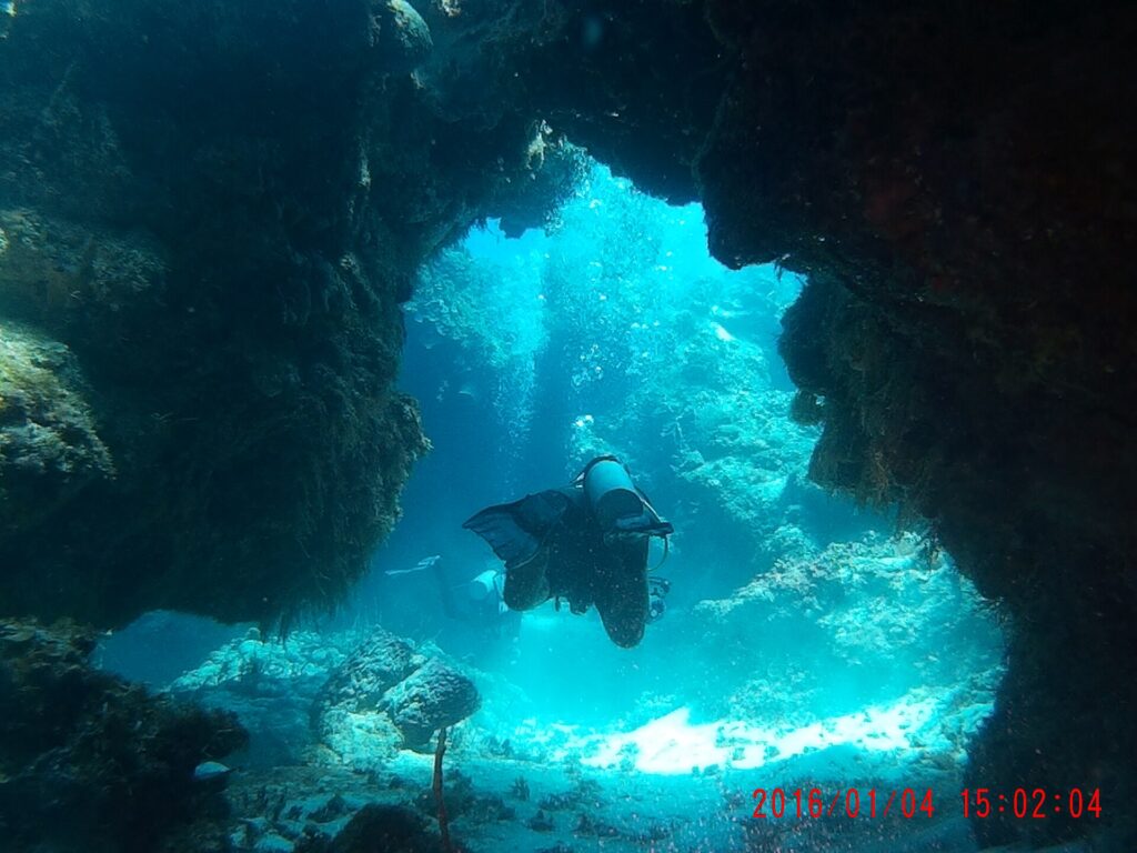A diver swims through a dark coral arch at Palancar, the white sand lit by the sun behind him