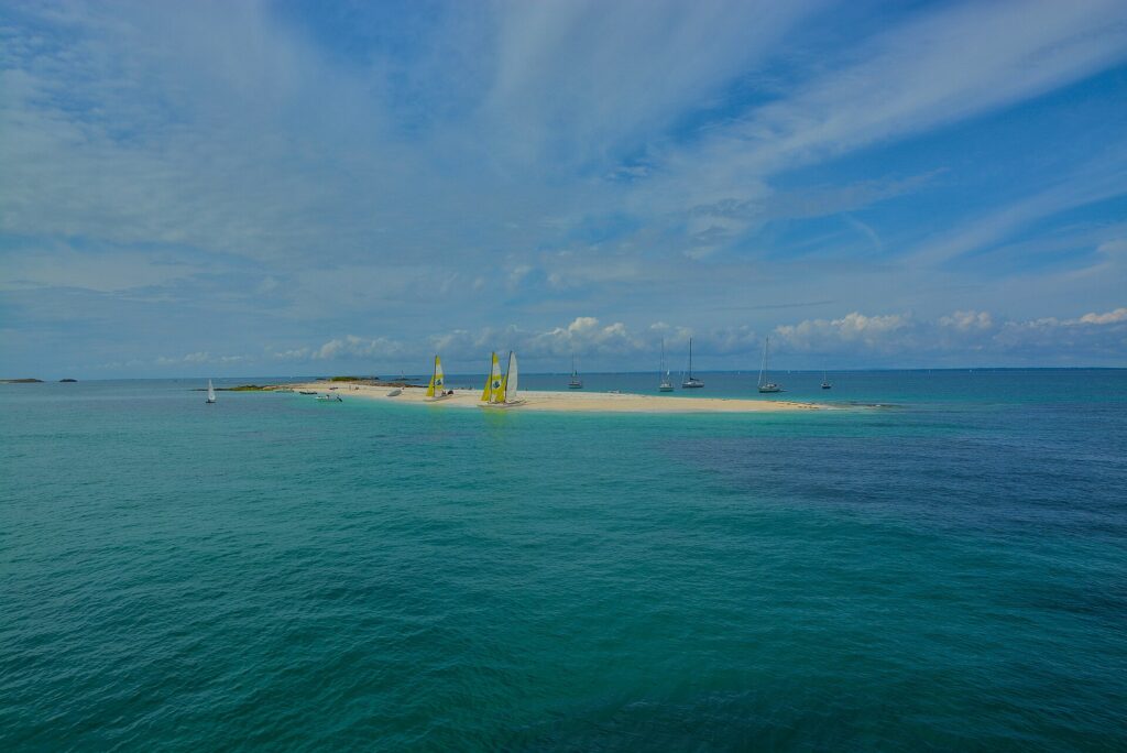 A long tongue of white sand where catamarans with yellow and white sails are beached, surrounded by sailboats at anchor on turquoise water, under a blue sky streaked with clouds.