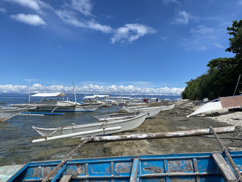 A row of white bangkas moored along a rocky shore, small outrigger canoes pulled up on the rock and a peeling blue boat in the foreground, under a blue sky with white clouds.
