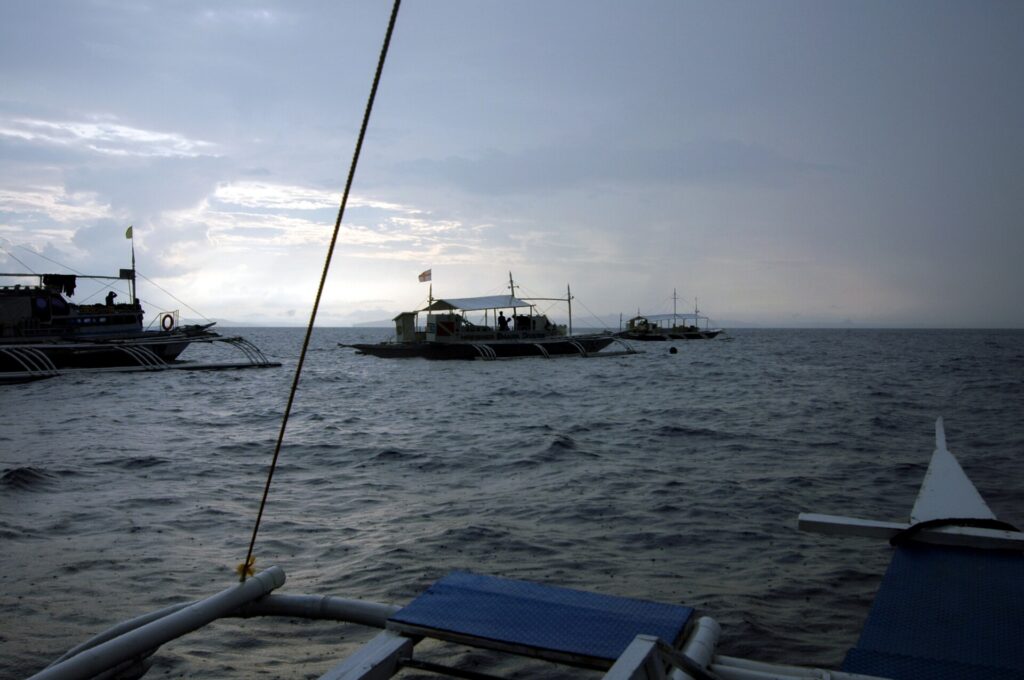 Three dive bancas moored on Monad Shoal under a gray early-morning sky, seen from the deck of another boat