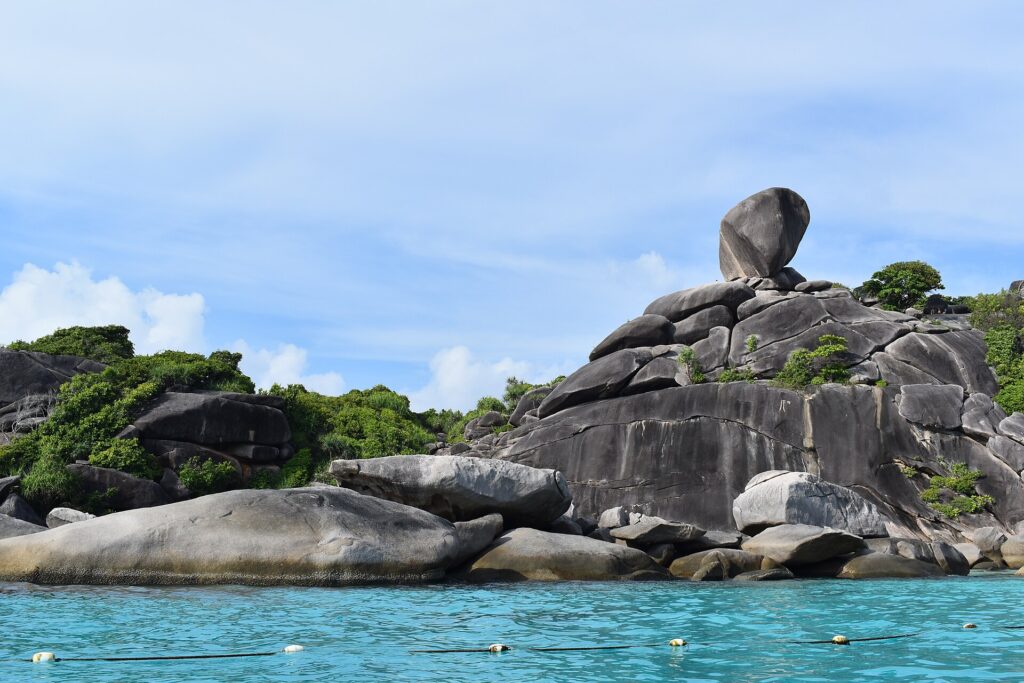 The huge balanced granite boulder of Island 8 tops a pile of smooth gray rocks at the edge of turquoise water, under a hazy blue sky, with a line of buoys in the foreground