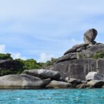The huge balanced granite boulder of Island 8 tops a pile of smooth gray rocks at the edge of turquoise water, under a hazy blue sky, with a line of buoys in the foreground