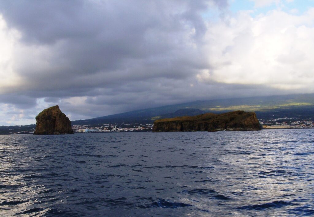 Seen from a boat on a dark, choppy sea, a tall rocky islet on the left and a long flat islet on the right, in front of the town of Madalena and the slopes of Pico under a heavy sky