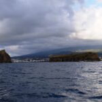 Seen from a boat on a dark, choppy sea, a tall rocky islet on the left and a long flat islet on the right, in front of the town of Madalena and the slopes of Pico under a heavy sky