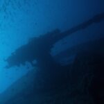 The stern gun of the Thistlegorm silhouetted against the blue, under a school of small fish