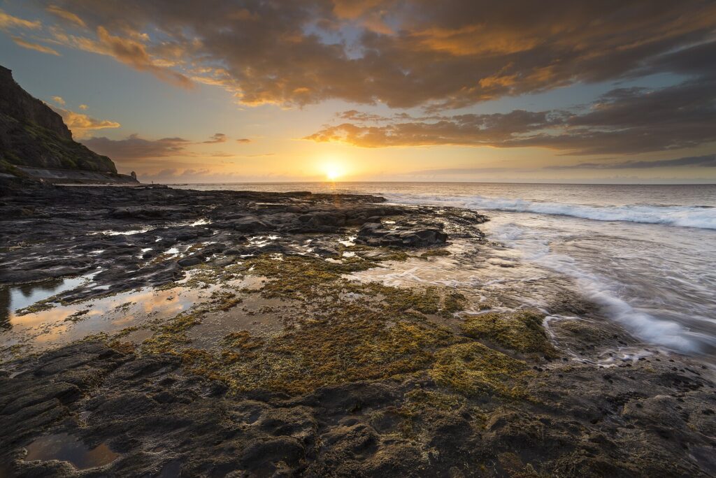 Sunset over the ocean seen from a black basalt rock platform covered in algae and tide pools, with waves lapping at it on the right and a cliff on the left
