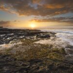 Sunset over the ocean seen from a black basalt rock platform covered in algae and tide pools, with waves lapping at it on the right and a cliff on the left