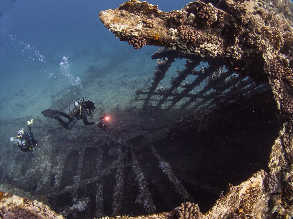 The open ribs of the Carnatic's hull, covered in coral, with two divers and a lit dive light inside