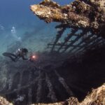 The open ribs of the Carnatic's hull, covered in coral, with two divers and a lit dive light inside