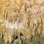 Close-up of pale yellow formations and thin stalactites, with the rippled reflection of the water below