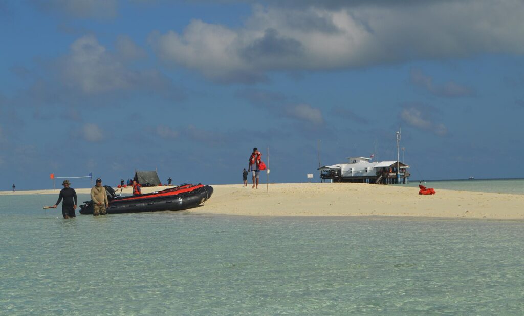 A white sandbank surrounded by clear pale green water, where a black and orange inflatable has just landed, people in life jackets on the sand and the ranger station on stilts on the right.