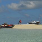 A white sandbank surrounded by clear pale green water, where a black and orange inflatable has just landed, people in life jackets on the sand and the ranger station on stilts on the right.