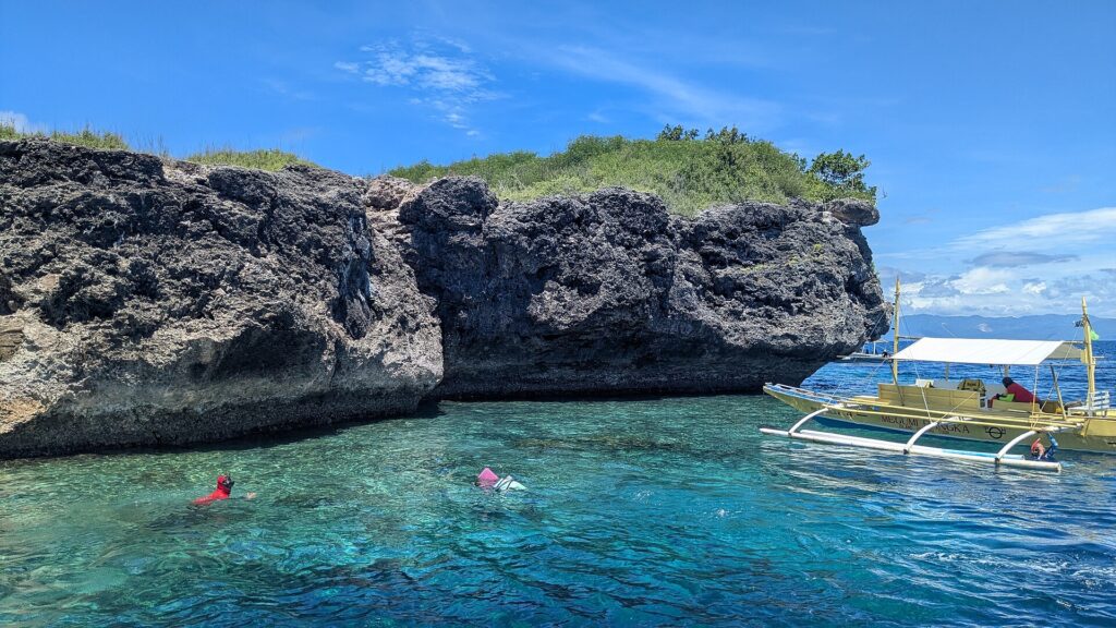 The gray, undercut limestone cliff of Pescador Island, two snorkelers and a yellow banca in turquoise water