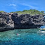 The gray, undercut limestone cliff of Pescador Island, two snorkelers and a yellow banca in turquoise water