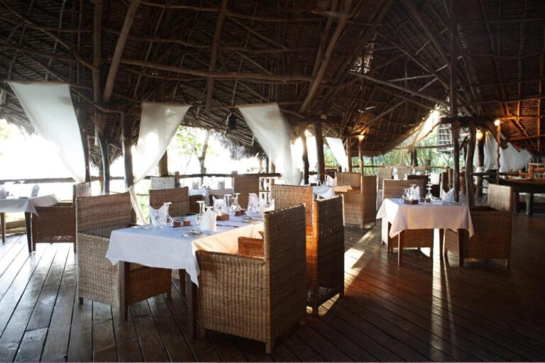 Tables with white tablecloths and wicker armchairs under a large thatched roof