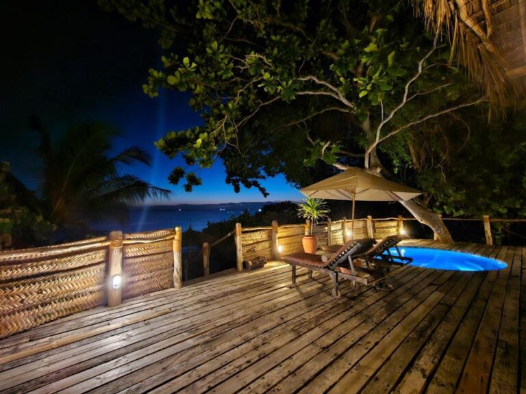 A wooden deck lit up in the evening, with a plunge pool, two sun loungers and a parasol