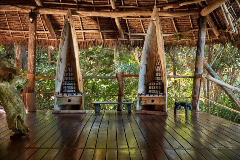 Two seats shaped like dugout canoes under a thatched roof, facing the greenery