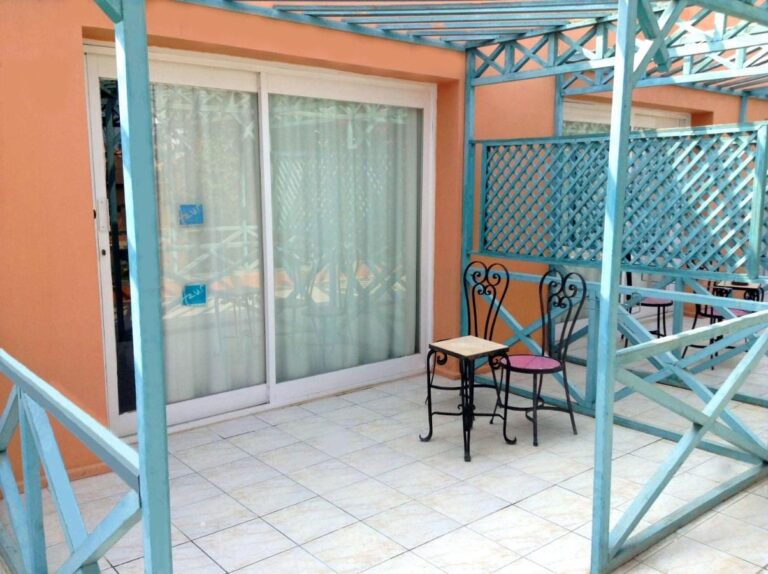 A tiled terrace under a turquoise pergola, with a small table and two wrought-iron chairs