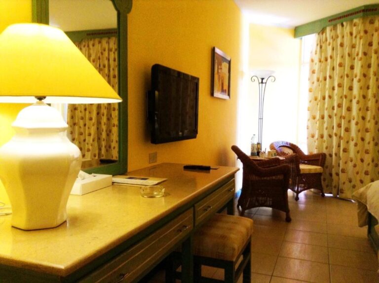 A room with yellow walls, a desk, a TV and two rattan armchairs by the window