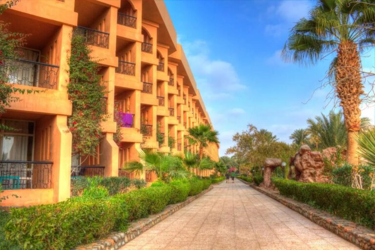 An ocher building with flower-filled balconies, along a path lined with hedges and palm trees