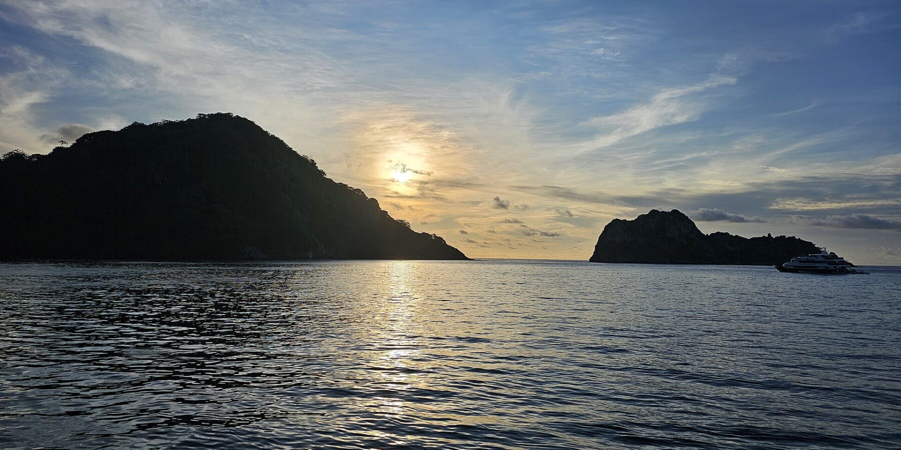 Cocos Island backlit at sunset, with a liveaboard at anchor on the right and a calm sea