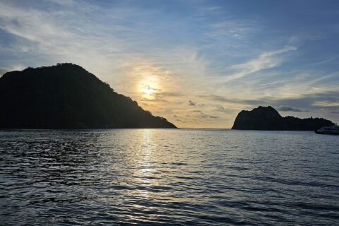 Cocos Island backlit at sunset, with a liveaboard at anchor on the right and a calm sea
