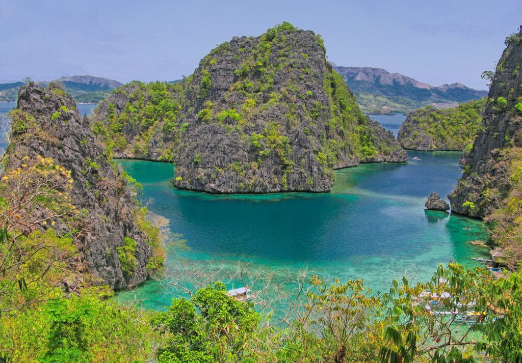 Looking down on a turquoise cove hemmed in by vegetated limestone peaks, a few bangkas moored below and hills on the horizon.