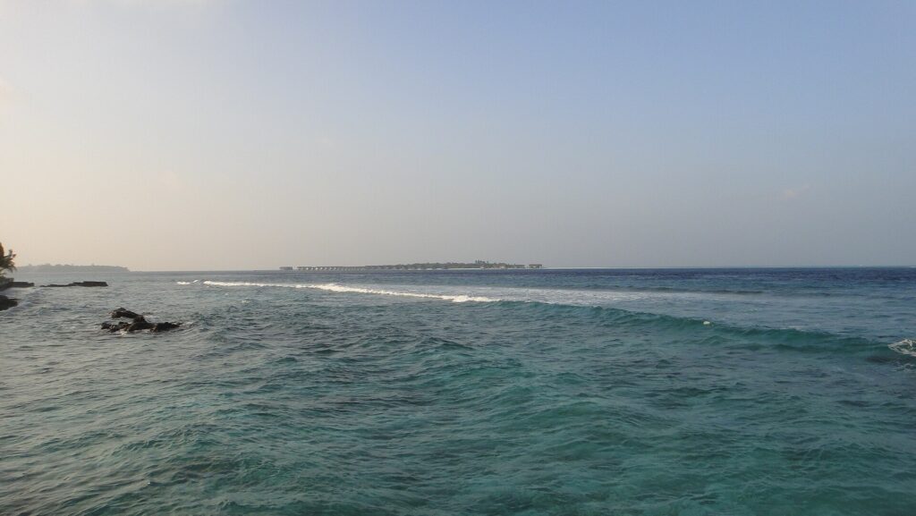 From a rocky shore, a choppy turquoise channel where a line of waves breaks on the reef, and on the horizon a low island lined with overwater bungalows, under a pale sky.