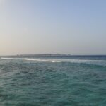 From a rocky shore, a choppy turquoise channel where a line of waves breaks on the reef, and on the horizon a low island lined with overwater bungalows, under a pale sky.