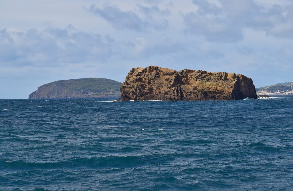 Ilhéu Deitado, a long block of ocher tuff with honeycombed cliffs, on a rough dark blue sea, with the green hill of Monte da Guia behind it and houses on Faial in the distance on the right