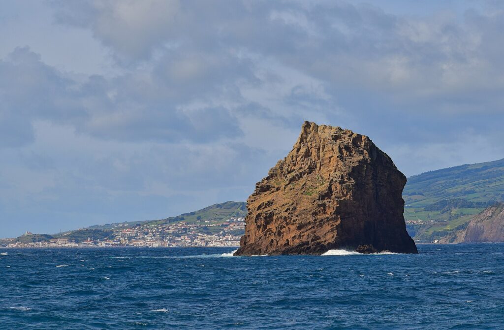 Ilhéu em Pé, a tall ocher tuff rock with eroded layers, rises from a dark blue sea where a few waves break, in front of the white town of Horta and the green hills of Faial under a cloudy sky