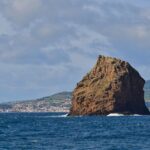 Ilhéu em Pé, a tall ocher tuff rock with eroded layers, rises from a dark blue sea where a few waves break, in front of the white town of Horta and the green hills of Faial under a cloudy sky