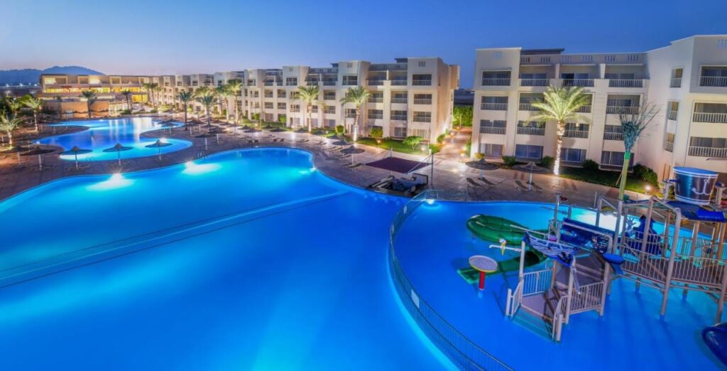The lit pools of JAZ Soma Beach at nightfall, between the buildings and the palm trees