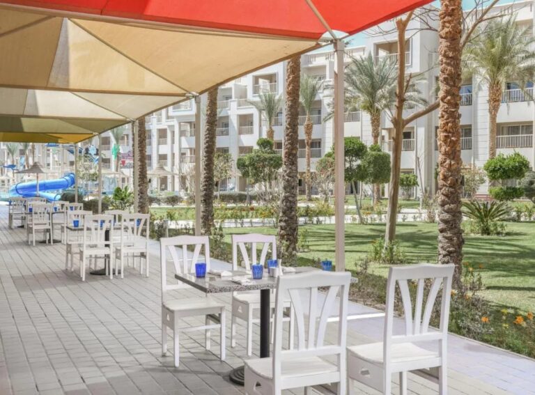 A restaurant terrace under shade sails, facing the garden and the palm trees
