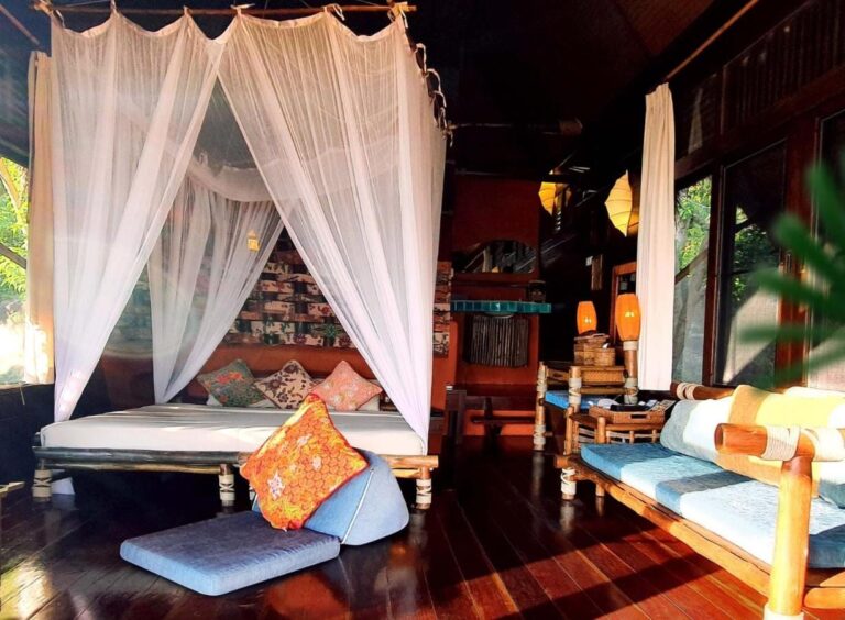A cottage with a bed under a mosquito net, a bamboo bench with blue cushions and a dark wooden floor