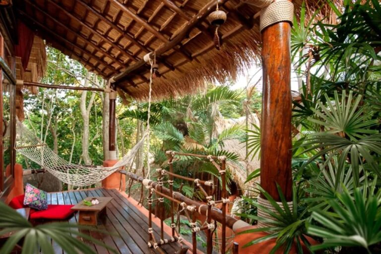 A wooden terrace with a hammock and red cushions, under a thatched roof, among the palm trees
