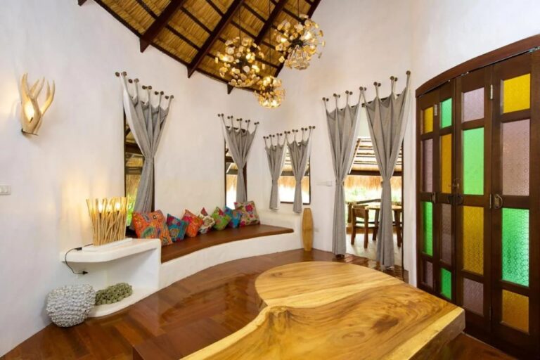 A white living room with a bench and colorful cushions, a solid wood table and stained-glass doors