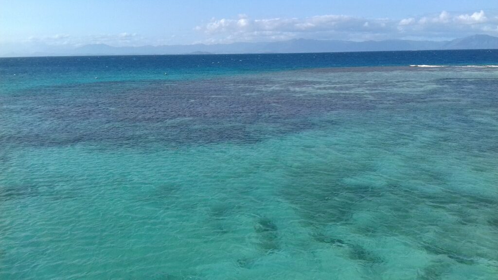 The lagoon from above, turquoise water dotted with dark coral heads, the barrier reef with breaking waves on the right and the mountains of Grande Terre on the horizon