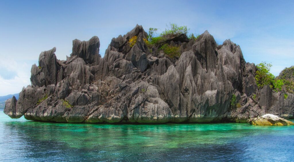 A spiky gray limestone islet topped with a little vegetation rises above clear turquoise water where coral shows through, under a blue sky.