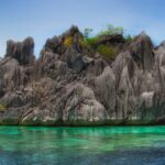 A spiky gray limestone islet topped with a little vegetation rises above clear turquoise water where coral shows through, under a blue sky.