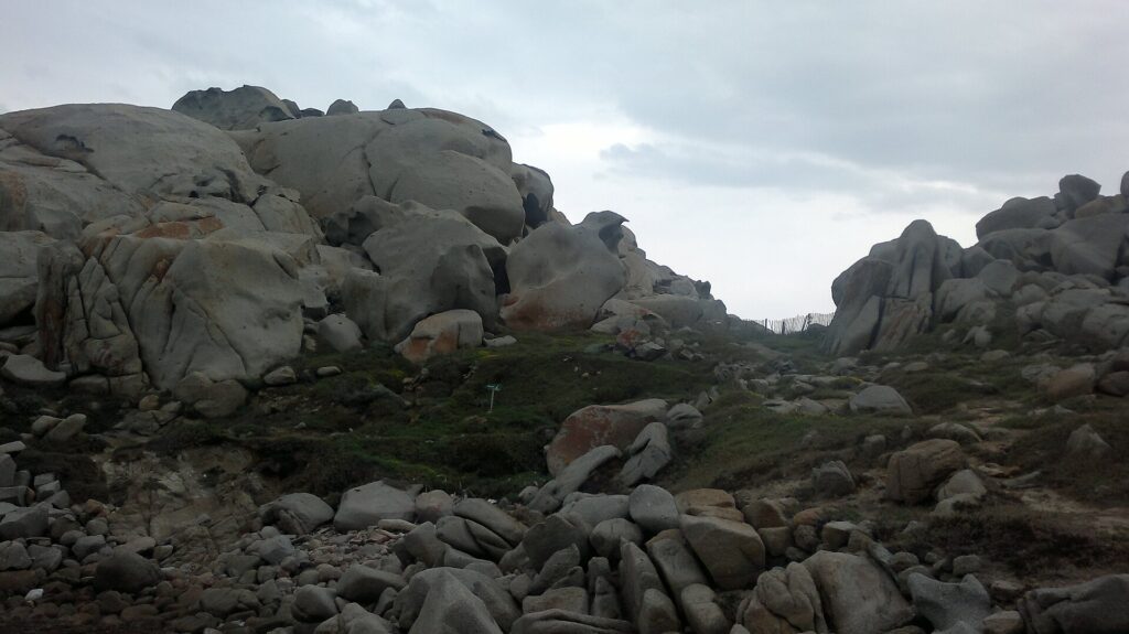 A jumble of large, rounded gray granite boulders hollowed by erosion and stained with orange lichen, around a short lawn where a path lined with a wooden fence runs toward the sea, under an overcast sky.