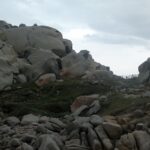 A jumble of large, rounded gray granite boulders hollowed by erosion and stained with orange lichen, around a short lawn where a path lined with a wooden fence runs toward the sea, under an overcast sky.