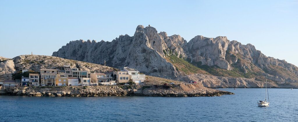 The houses of Les Goudes village lined up right along a rocky shore, in front of a high ridge of white limestone with jagged peaks, under a blue sky, with a sailboat at anchor on a calm sea.