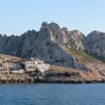The houses of Les Goudes village lined up right along a rocky shore, in front of a high ridge of white limestone with jagged peaks, under a blue sky, with a sailboat at anchor on a calm sea.