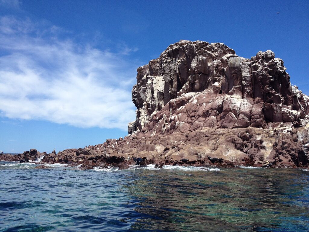 The reddish rocks of Los Islotes under a blue sky, with sea lions lying at the waterline and a cracked cliff above