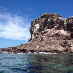 The reddish rocks of Los Islotes under a blue sky, with sea lions lying at the waterline and a cracked cliff above