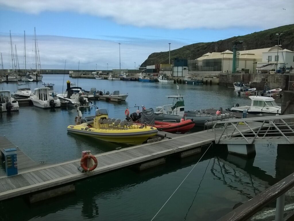 RIBs, a yellow one with rows of seats and a red one, moored at the Vila do Porto marina pontoon, with sailboats and fishing boats behind under a green cliff and a cloudy sky