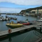 RIBs, a yellow one with rows of seats and a red one, moored at the Vila do Porto marina pontoon, with sailboats and fishing boats behind under a green cliff and a cloudy sky