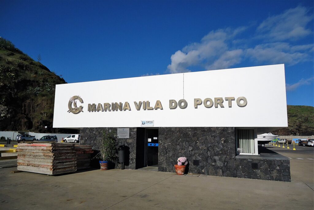 The reception building of the Vila do Porto marina, a large white sign with metal letters on a black basalt wall, under a blue sky, with a green cliff on the left